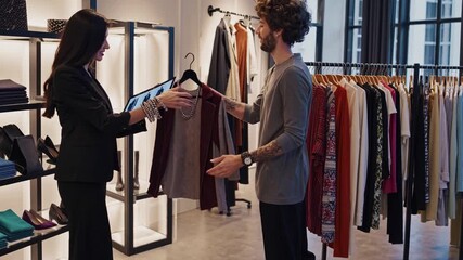 Boutique stylist advising customer with tablet in modern clothing store, racks of dresses and shoes, male customer examining garment, sales consultation, digital lookbook, warm lighting, polished - Powered by Adobe
