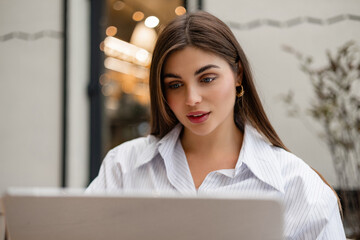 Close-up of young woman looking at laptop screen while working at outdoor cafe. Focused freelancer using computer for business or studying, modern lifestyle concept.