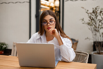 Young woman in glasses working on laptop at outdoor cafe. Focused freelancer using computer for business or studying, modern technology and remote work concept.