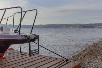 Small motorboat on a wooden platform, gentle water, and a pebbled shoreline with distant hills. Travel, nature, and outdoor leisure.