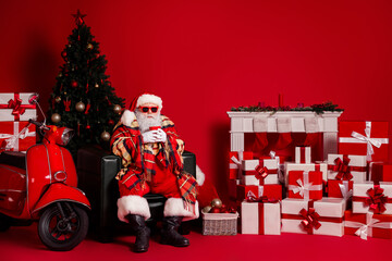 Santa sits in a red room with a coffee mug surrounded by gifts a Christmas tree and a red scooter