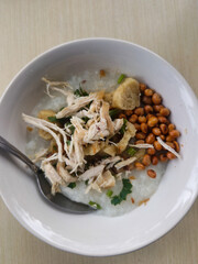 A delicious bowl of Indonesian chicken porridge, known as Bubur Ayam, topped with shredded chicken, roasted peanuts, and fresh herbs, served with a spoon.