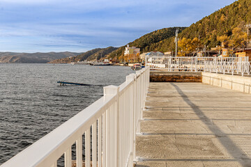 A promenade with white railings along Lake Baikal and surrounding mountains by Listvyanka Village where the Angara River begins in autumn on a sunny day.
