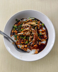 A white bowl of Indonesian chicken porridge (bubur ayam) topped with shredded chicken, peanuts, and fried shallots on a wooden table.
