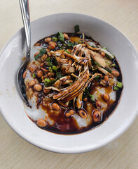 A bowl of traditional Indonesian chicken porridge, Bubur Ayam, garnished with shredded chicken, roasted peanuts, and green onions, served with a spoon.