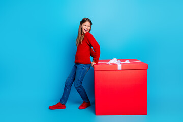 Girl in red sweater stands beside a large red gift box against a bright blue backdrop for festive...