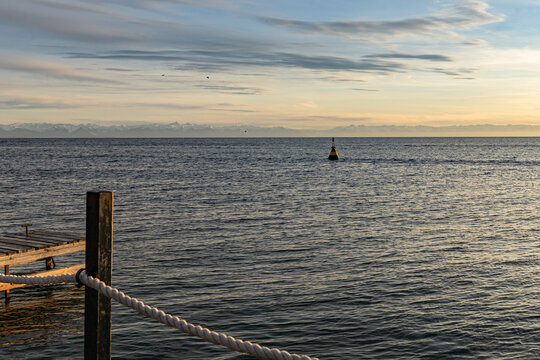 A tranquil coastal scene at dusk with a weathered wooden pier with a rope railing, calm waters, and a single buoy on the open sea under a soft, colorful sky. - Powered by Adobe