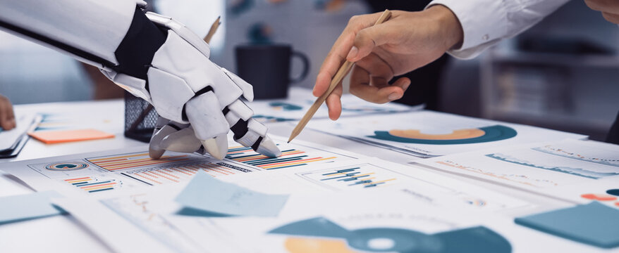 A modern office scene showcasing collaboration between a robotic hand and a human hand, analyzing various data charts and graphs for innovative business solutions.Noogenesis - Powered by Adobe