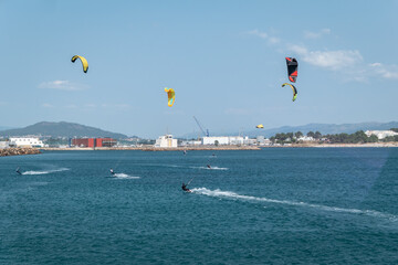 Alguns kitesurfistas a deslizarem sobre as águas azuis-turquesa junto à costa, com as suas velas coloridas erguidas contra um céu azul em Portugal