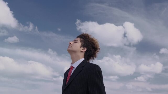 Portrait of a handsome teenager in a business suit against a backdrop of a hill and blue sky, his face upturned and his curls fluttering in the wind. The camera pans