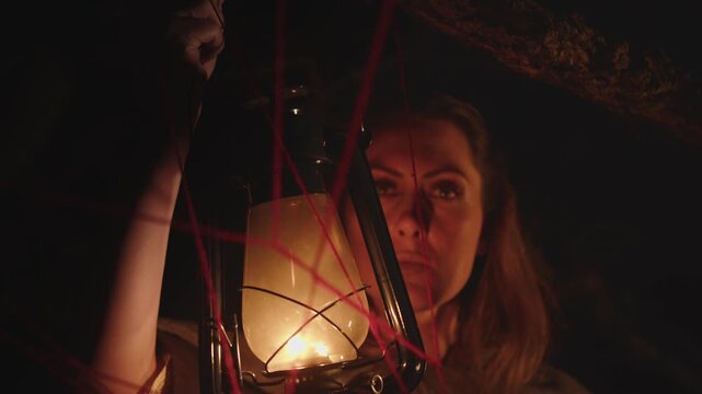 A woman looks at a chaotic interweaving of red threads in the dark and her face is illuminated by the flickering light of a kerosene lamp