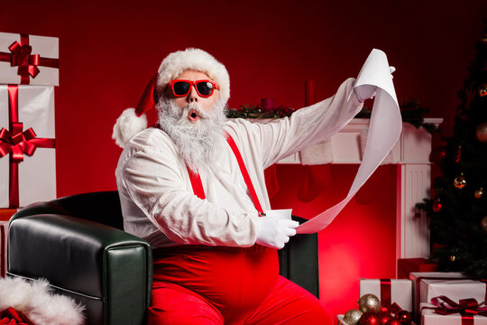 Santa with red suspenders and white beard reads a long list while posing with gifts by the Christmas tree and cozy fireplace - Powered by Adobe