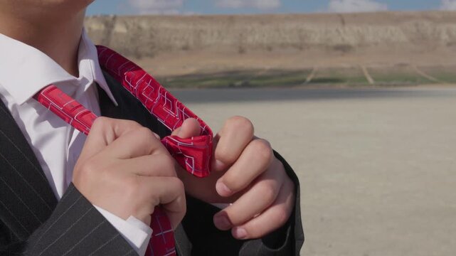A curly-haired teenager with glasses and a mature business suit unties and removes his tie against a backdrop of a hill and blue sky. The camera is moving.
