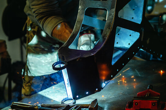 View through metal parts covering device, master welds parts. Yellow sparks, smoke. Welding workshop