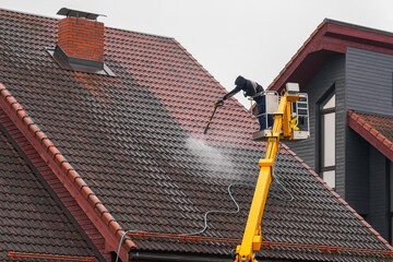 Worker cleaning house roof with high-pressure washer from lift platform on a rainy day