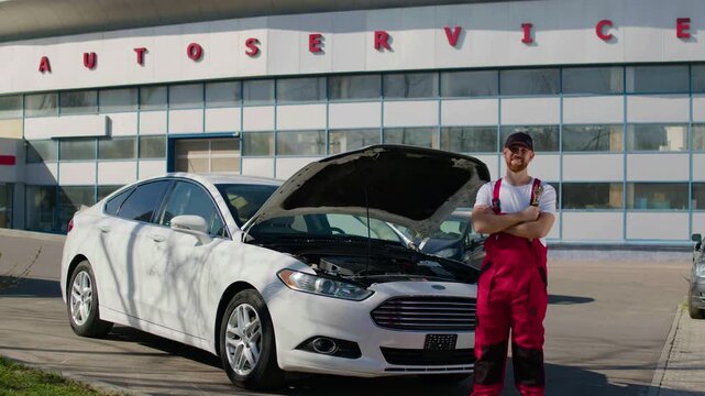 Male auto technician in red overalls stands confidently beside a white car with an open hood, showcasing expertise in automotive diagnostics and repair services