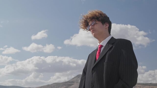 Curly-haired teenager in glasses and a business suit with a red tie against the backdrop of a mountain landscape and blue sky