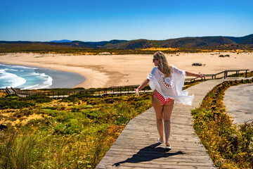 Visiting Algarve coast in Portugal. Beautiful middle-aged woman walking on boardwalk to sandy Bordeira beach and enjoying vacations over ocean on summer day. Back view