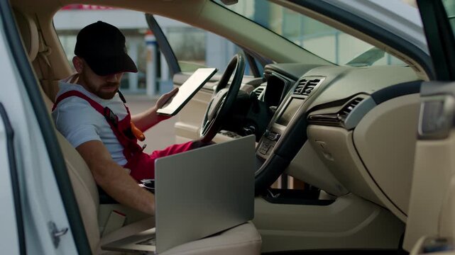 Mechanic in red overalls, seated inside a modern vehicle, using a tablet to discuss vehicle diagnostics and repair solutions with a customer in a professional setting