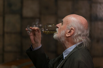 An elderly man savors a glass of wine, appreciating its aroma and taste at a wine tasting event indoors.