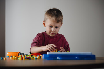 A young boy is carefully assembling a vibrant model using building blocks. He holds a green tool and shows concentration, working quietly at a wooden table in a bright room.