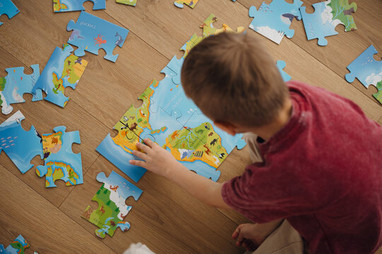 A young child focuses on completing a colorful puzzle featuring a world map. Surrounding pieces are scattered on a wooden floor, creating a playful learning environment. top view