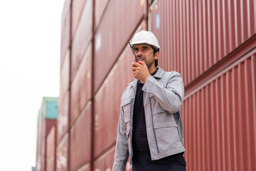 Male engineer in safety helmet using walkie talkie while inspecting stacked cargo containers at an outdoor logistics terminal shipping yard.