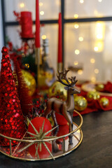 Beautiful Christmas composition with red decorative trees, a golden reindeer figurine, and shiny gold ornaments on a golden tray. In the background — a glowing Christmas tree with warm fairy lights. 