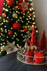 Beautiful Christmas composition with red decorative trees, a golden reindeer figurine, and shiny gold ornaments on a golden tray. In the background — a glowing Christmas tree with warm fairy lights. 