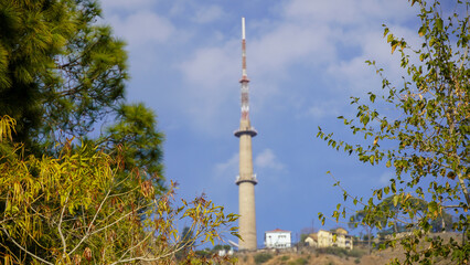 Fototapeta premium A tall, concrete communication tower with red & white striped markings stands prominently on a hill against a clear blue sky with white clouds. Lush green foliage & trees in the foreground in kasauli.