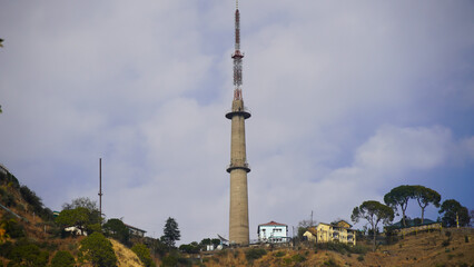 A tall, concrete communication tower with red & white sections stands on a hillside under a...