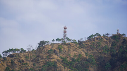A large, concrete communication tower with multiple tiers & red/white markings stands prominently on a hilltop under a cloudy sky. Residential buildings & trees are visible near the base of the tower.