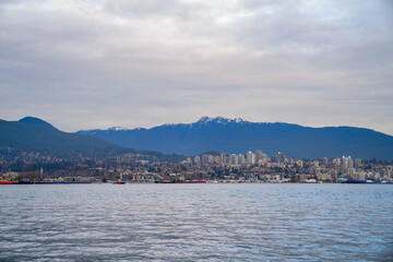 Fototapeta premium A panoramic view of a coastal city beneath expansive mountains, with shoreline homes, towers, and calm harbor water forming a balanced scenic landscape.