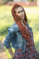 Naklejka premium Young woman with long red hair wearing floral dress and denim jacket poses outdoors, showcasing vibrant style and playful attitude in a natural setting