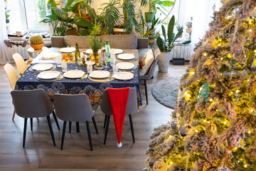 A festive table set for guests for Christmas and New Year, a Christmas tree in the living room, plates, bottles, glasses, potted plants, fairy lights