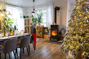 Festive interior of house is decorated for Christmas and New Year in loft style with black stove, fireplace, Christmas tree. Warm studio room with set table, burning wood, cozy and heating of home