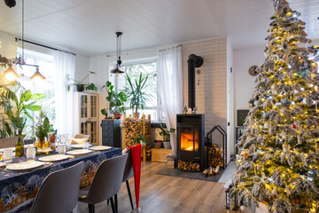 Festive interior of house is decorated for Christmas and New Year in loft style with black stove, fireplace, Christmas tree. Warm studio room with set table, burning wood, cozy and heating of home