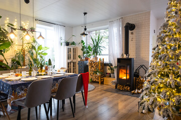 Festive interior of house is decorated for Christmas and New Year in loft style with black stove, fireplace, Christmas tree. Warm studio room with set table, burning wood, cozy and heating of home