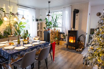 Festive interior of house is decorated for Christmas and New Year in loft style with black stove, fireplace, Christmas tree. Warm studio room with set table, burning wood, cozy and heating of home