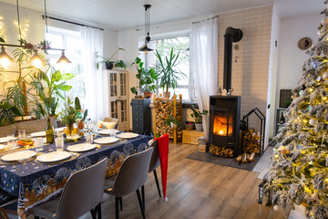 Festive interior of house is decorated for Christmas and New Year in loft style with black stove, fireplace, Christmas tree. Warm studio room with set table, burning wood, cozy and heating of home