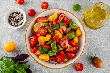 A salad of farm-fresh multi-colored tomatoes, red onions and fresh basil in a bowl on a gray concrete background. Healthy food. Top view.