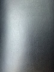 Close-up of a dark gray or black faux leather surface with a detailed texture. A light gradient across the material highlights its rough grain. Used as a background, textile, design, and fashion.