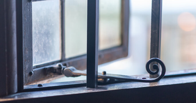 Close-up of an old, dark metal window frame with an ornate scroll latch. Historical architectural detail with blurred bright light for vintage, contemplation, or interior design concepts.