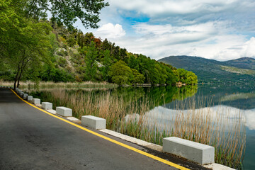 Fototapeta premium Scenic asphalt road along Lake Orestiada in Kastoria, northern Greece, with reeds, lush green hills, forest reflections, and dramatic cloudy sky creating a tranquil travel destination in the Balkans.