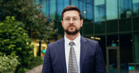 Fototapeta premium Portrait of serious and successful Caucasian businessman in suit and glasses, standing on the street in front of modern office building, confidently looking at the camera headshot of corporate manager