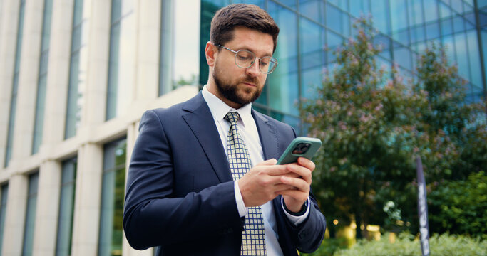 Confident Caucasian man in jacket and tie walking outdoors on a sunny summer day, using a smartphone and typing a message with a modern glass building behind him