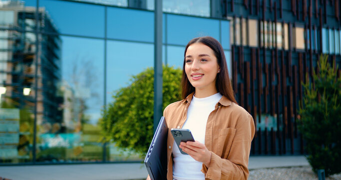 A beautiful young woman walks by a commercial building while texting on her smartphone and holding laptop yet professional appearance. Urban background. Modern lifestyle, freelance or business concept