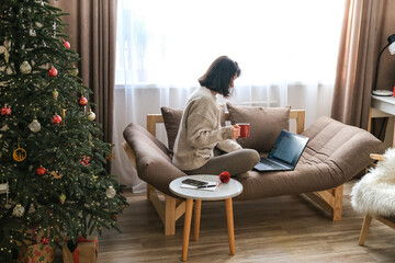 Woman sits on sofa with Christmas tree nearby, working on laptop and holding red mug, creating cozy holiday work-from-home scene. Remote work, online shopping, freelance work during festive period
