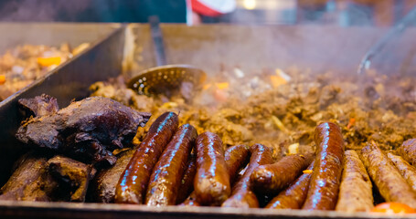 Street vendor grilling variety of sausages and meats on sizzling hot plate, using skimmer during busy market food preparation