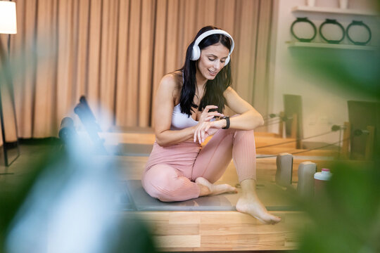 A focused woman seated cross-legged checks her smartwatch while enjoying her workout playlist, radiating concentration and modern fitness lifestyle in a serene environment.